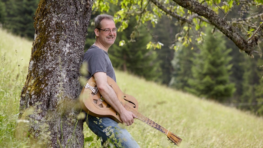 Der Musiker Gottfried Gfrerer lehnt mit seiner Resonatorgitarre an einem Baum.